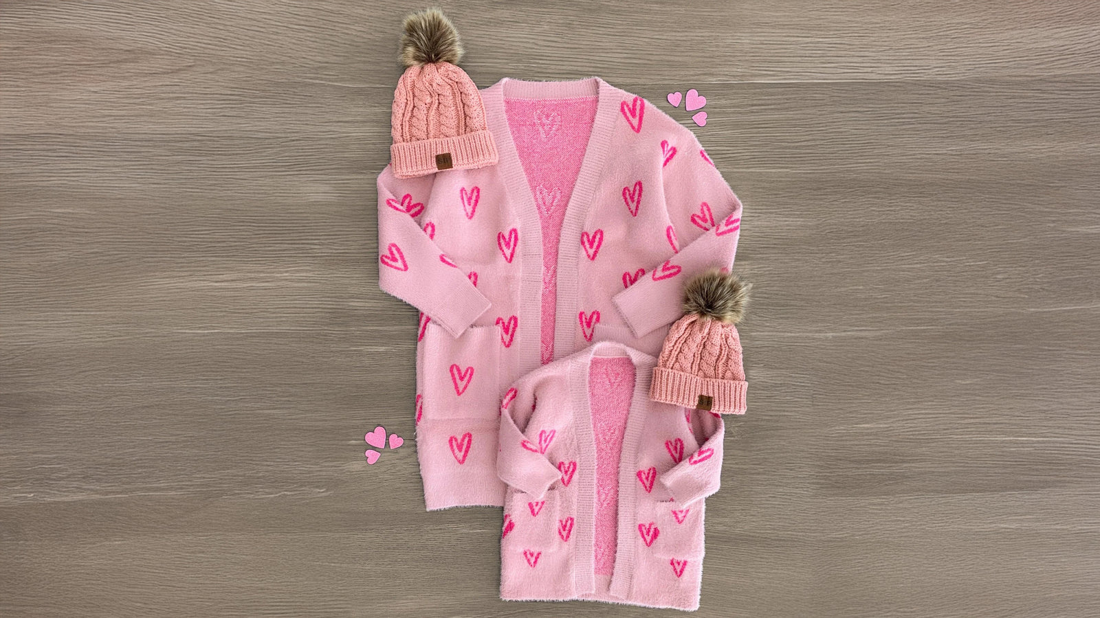 Two matching pink heart cardigans, one for mom and one for her little one, laid flat to showcase their long sleeves and light pink fabric with bright hot pink heart patterns. Made from soft and cozy material, these cardigans are styled for comfort and versatility. Perfect for pairing with Sparkle in Pink (SIP) bottoms and booties, creating an adorable twinning look for mommy and her mini BFF