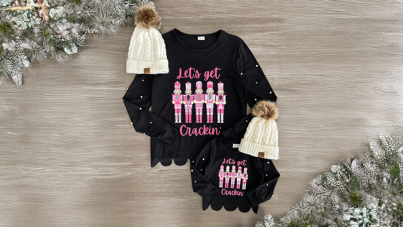 Matching mom and child long sleeve black tops featuring pearl details, a scalloped hem, and a pink nutcracker design with 'Let's Get Crackin'' on the front. Made from soft, breathable fabric, these tops provide comfort for all-day wear. Perfect for a coordinated, festive look, they can be paired with various bottoms and booties for holiday adventures.